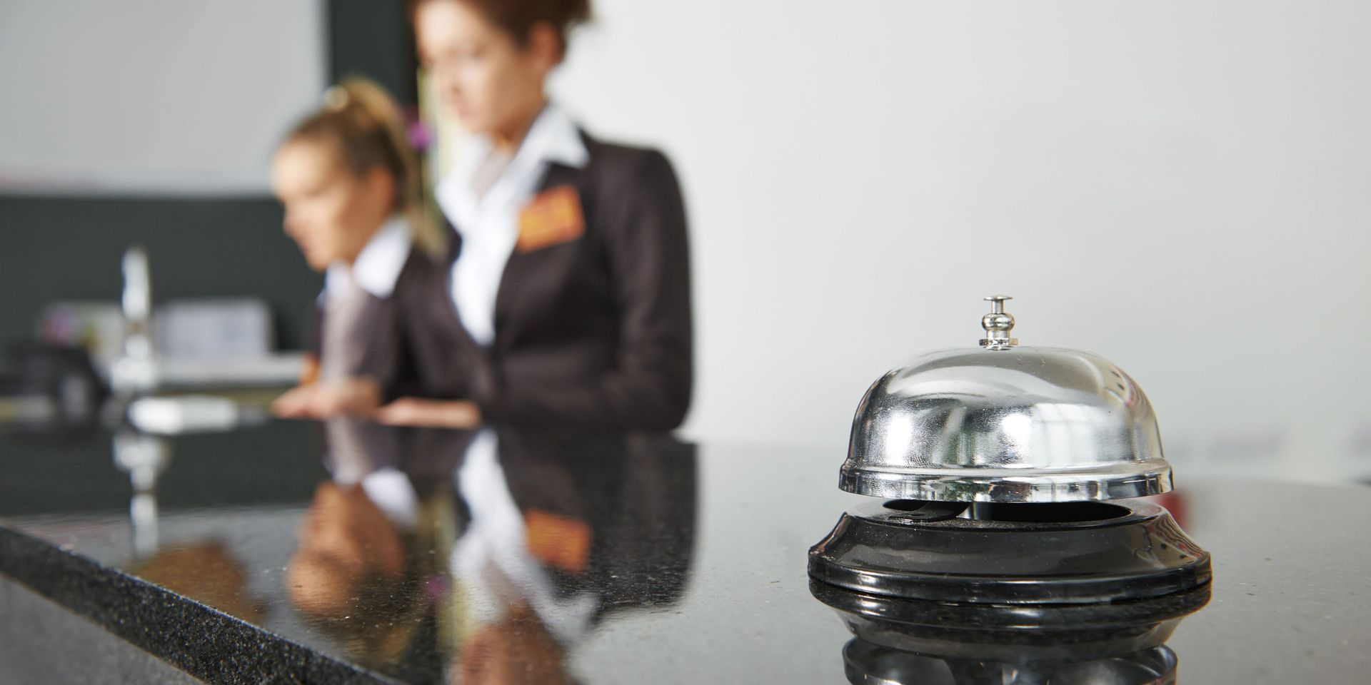 Recepcionistas de hotel uniformizadas atrás do balcão, com a sineta em primeiro plano
